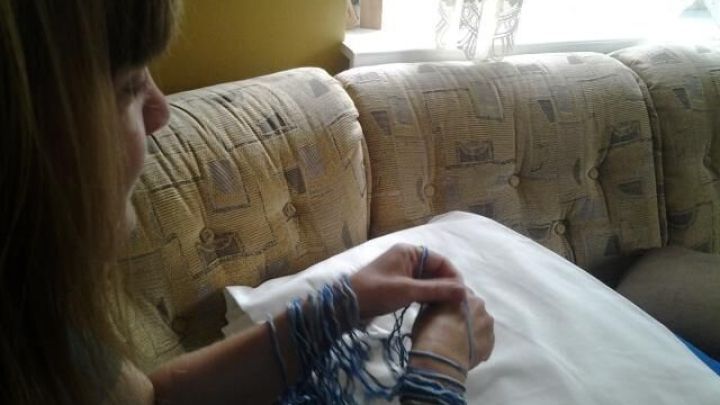 Eine Person strickt mit blauer Wolle ohne Nadeln, entspannt auf einer Couch im Wohnzimmer mit hellem Licht.
