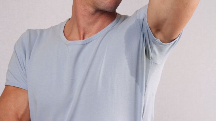 Ein Mann hebt seinen Arm, w&auml;hrend schwei&szlig;fleckige Stellen auf seinem hellblauen T-Shirt sichtbar sind.