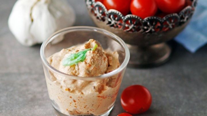 Ein Tomaten-Dip in einem Glas steht neben einer Schale mit Kirschtomaten und einer Knoblauchknolle auf einem Tisch.