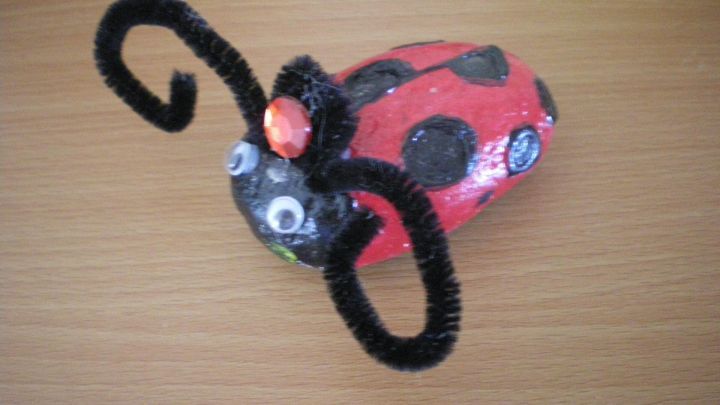 Ein bunter K&auml;fer aus einem Stein mit Antennen aus Pipe Cleaners liegt auf einem Holzuntergrund und erweckt Freude.
