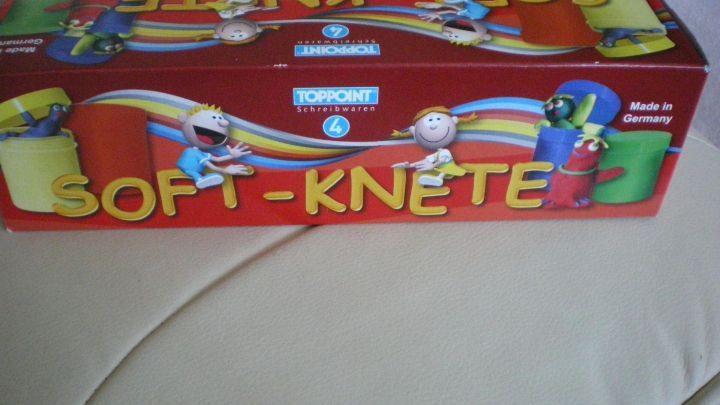 Eine rote Schachtel Soft-Knete liegt auf einem Sofa. Auf der Schachtel steht: "TOPPOINT Schreibwaren 4, Made in Germany."