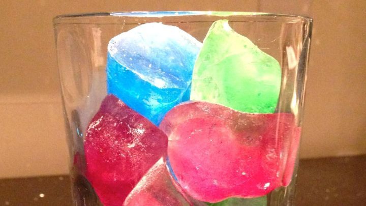 Bunte Eisw&uuml;rfel liegen in einem Glas, w&auml;hrend leuchtend rote, gr&uuml;ne und blaue St&uuml;cke auf einer dunklen Oberfl&auml;che verstreut sind.