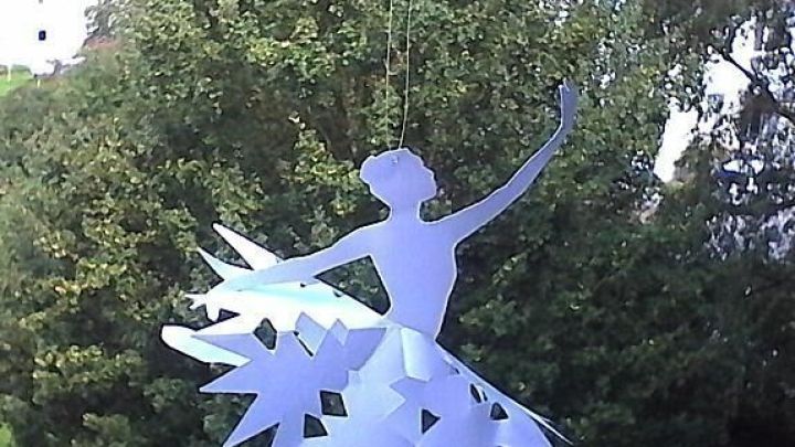 Papier-Ballerina h&auml;ngt elegant vor &uuml;ppigem, gr&uuml;nem Hintergrund und dient als stilvolle Dekoration.
