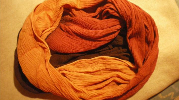 Ein bunter Schal aus warmen Farbt&ouml;nen liegt verdreht in Loop-Form auf einer beige Oberfl&auml;che.