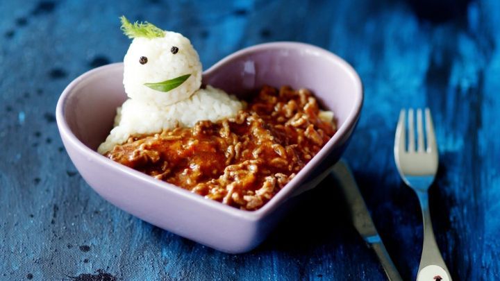 Ein herzf&ouml;rmiger Teller mit Reis, dekoriert mit einem l&auml;chelnden Reismann aus Reis und Kr&auml;utern, auf einem blauen Untergrund.