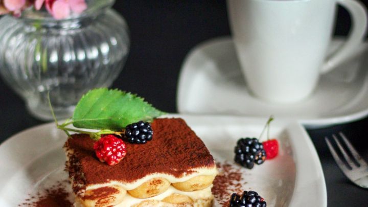 Eine k&ouml;stliche, kalorienarme Tiramisu-Scheibe liegt auf einem wei&szlig;en Teller, umgeben von Beeren und einem Blumenstrau&szlig;.