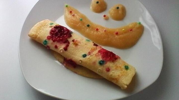Eingerollte Pfannkuchen mit Himbeeren und bunten Zuckerperlen liegen auf einem Teller, umgeben von Fruchtp&uuml;ree mit Smiley-Gesicht.