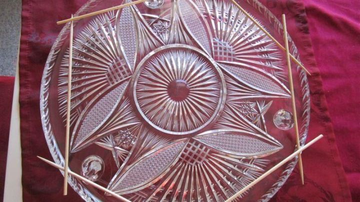 Ein gl&auml;serner Tortenteller mit einem dekorativen Muster ist auf einem roten Tisch liegend, stabilisiert mit Holzst&auml;bchen.