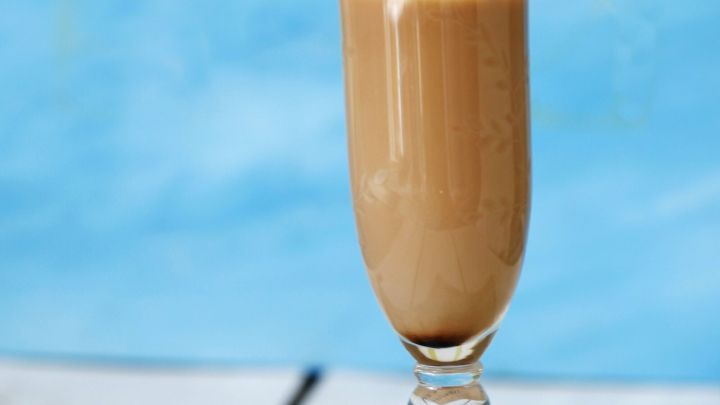 Ein elegantes Glas mit Kaffee und wei&szlig;em Rum steht auf einem Tisch, dekoriert mit einer Orangenscheibe und einem Strohhalm. Hintergrund in sanften Blaut&ouml;nen.