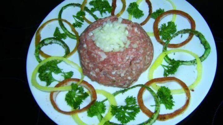 Auf einem wei&szlig;en Teller sind farbige Zwiebelringe um eine Form aus rohem Hackfleisch angeordnet, garniert mit frischem Koriander.