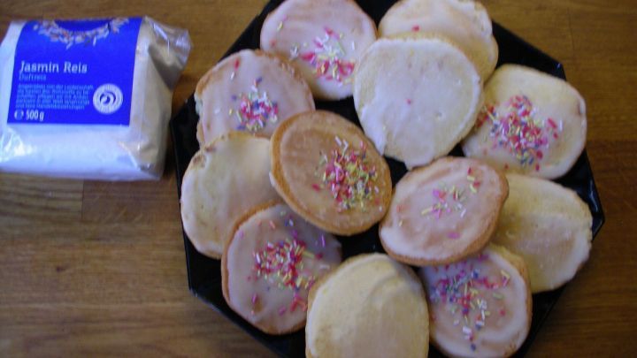 Auf einem Holzuntergrund liegen Kekse mit Zuckerguss und bunten Streuseln, daneben steht ein Beutel mit Reismehl.