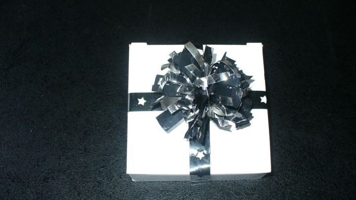 Ein wei&szlig;es Geschenk mit schwarzem Band und einer Schleife liegt auf einem schwarzen Tisch, passend zur Deko aus Musik- und Videokassetten.