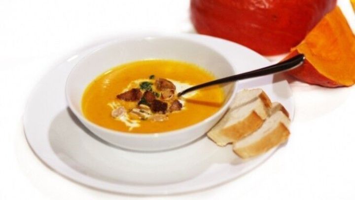 Eine Schale K&uuml;rbissuppe mit Croutons und Sahne, daneben Brotscheiben, ruht auf einem wei&szlig;en Teller. Im Hintergrund ein K&uuml;rbis.