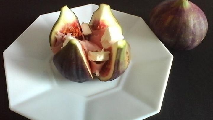 Gebackene Feigen sind auf einem wei&szlig;en Teller aufgeschnitten und gef&uuml;llt mit K&auml;se und Schinken, daneben liegt eine ganze Feige.