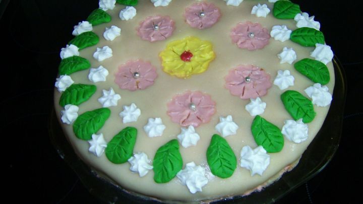 Eine runde Marzipandecke mit bunten Blumen und gr&uuml;nen Bl&auml;ttern dekoriert, auf einem glatten Untergrund pr&auml;sentiert.