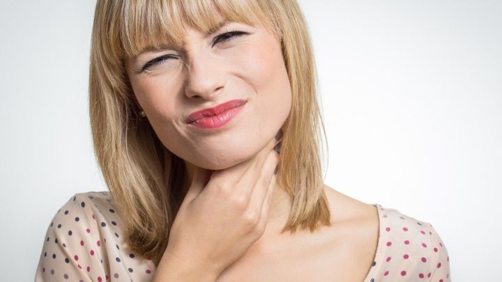 Eine Frau fasst sich mit einer Hand an den Hals und zeigt ein schmerzhaftes Gesicht, w&auml;hrend sie an Rachenbeschwerden leidet.