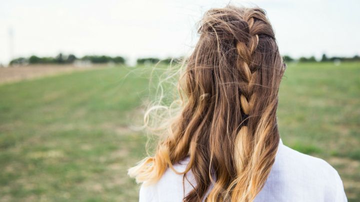 Lange, leicht gewellte Haare mit Zopf wehen im Wind auf einem offenen Feld unter einem bew&ouml;lkten Himmel.