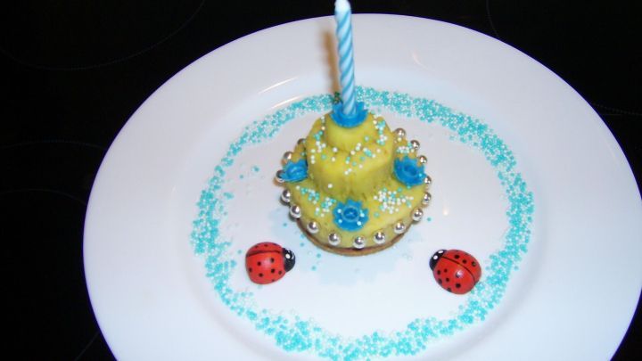 Gelbes Mini-Marzipan-T&ouml;rtchen mit Kerze und bunten Streuseln, dekoriert auf einem wei&szlig;en Teller mit blauen Zuckerperlen.