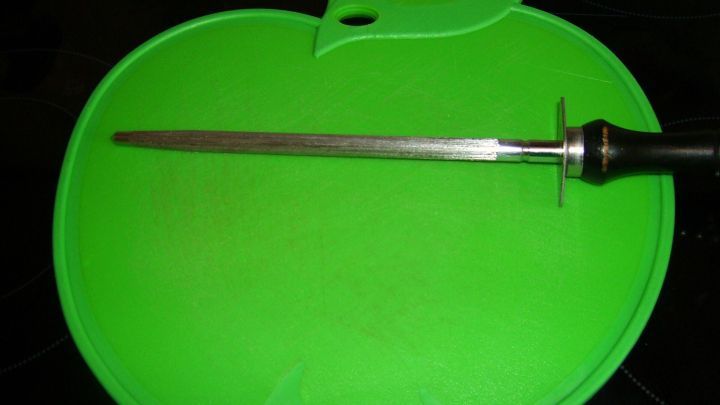 Ein gr&uuml;nes Schneidebrett in Apfelform liegt auf einer dunklen Oberfl&auml;che, w&auml;hrend ein langes Messer darauf platziert ist.