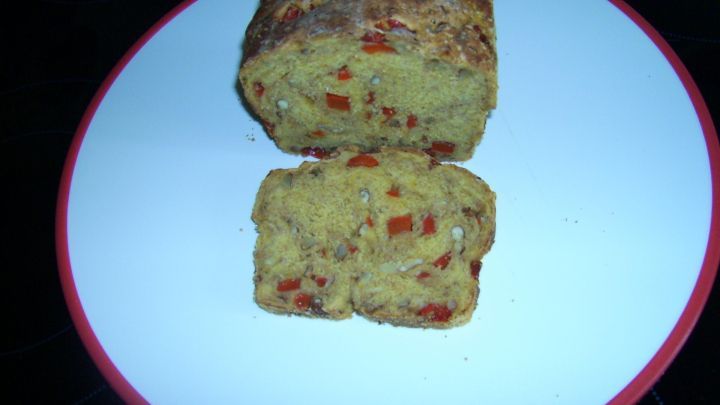 Ein golden gebr&auml;untes K&uuml;rbisbrot liegt auf einem wei&szlig;en Teller, mit einer Scheibe daneben, in einem schlichten, dunklen Umfeld.