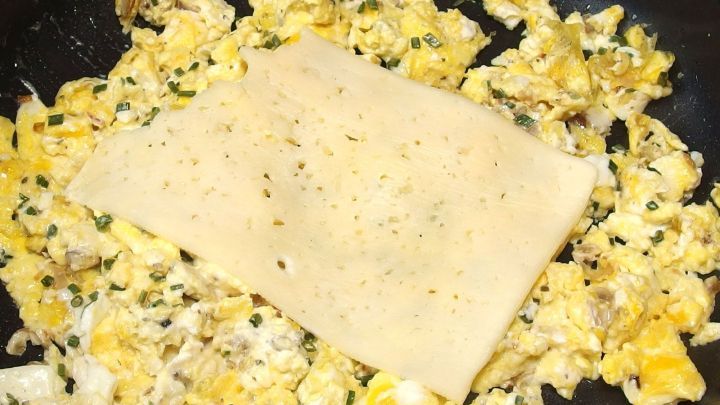 R&uuml;hrei mit K&auml;se wird in einer Pfanne zubereitet, w&auml;hrend eine K&auml;sescheibe darauf schmilzt und das Gericht verfeinert.