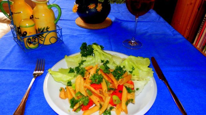 Eine Teller Nudeln liegt auf einem blauen Tisch, umgeben von frischem Gem&uuml;se und einem Glas Wein, bereit zum Essen.