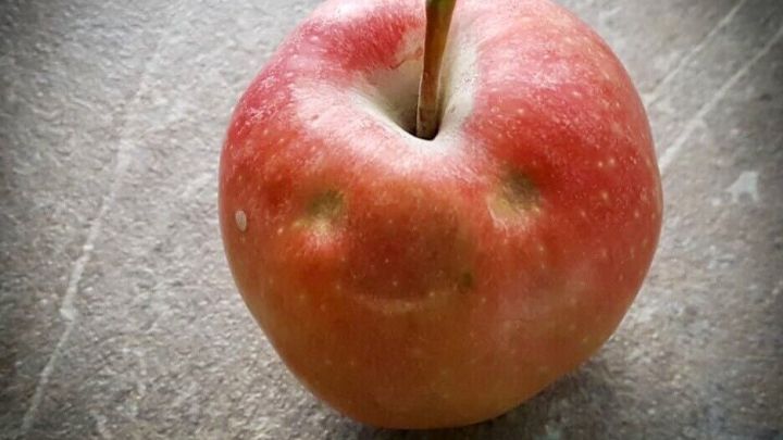 Ein frischer, roter Apfel mit einem l&auml;chelnden Gesicht steht auf einer grauen, strukturierten Oberfl&auml;che und symbolisiert gesundes Abnehmen.