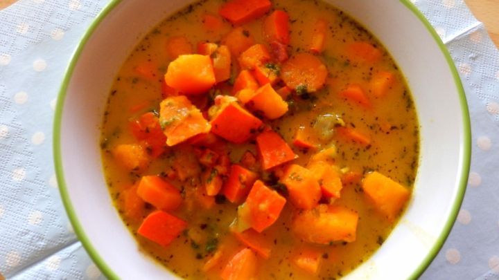 Eine cremige K&uuml;rbis-M&ouml;hren-Suppe mit bunten Gem&uuml;sest&uuml;cken in einer wei&szlig;en Sch&uuml;ssel, auf einem blauen Untergrund.