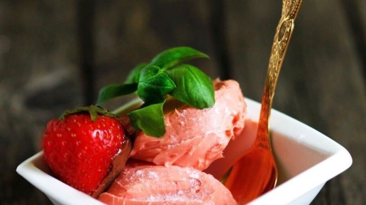 Eine wei&szlig;e Sch&uuml;ssel mit Erdbeereis, garniert mit einer Erdbeere und frischem Basilikum, steht auf einer rustikalen Holzoberfl&auml;che.