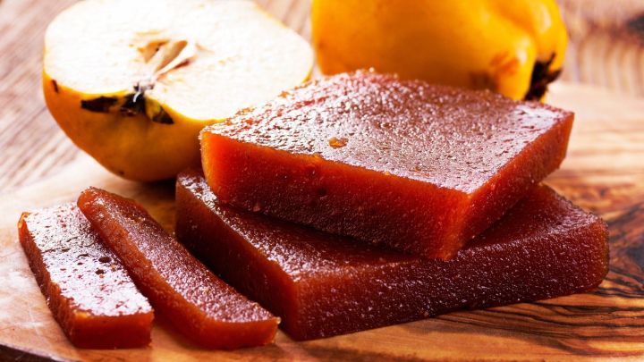 Dicke, gl&auml;nzende Quittenbrotst&uuml;cke liegen auf einem Holzbrett, w&auml;hrend eine frische Quitte daneben liegt, umgeben von nat&uuml;rlichem Licht.