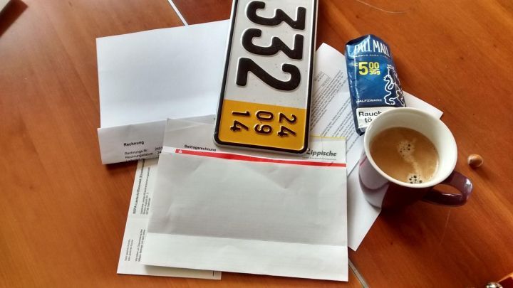 Ein Stapel diverser Briefe und Dokumente liegt auf einem Tisch, daneben eine Tasse Kaffee und ein Nummernschild.