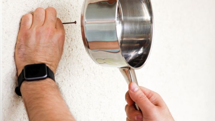 Eine Hand h&auml;lt einen Kochtopf, der als Hammerersatz verwendet wird, um einen Nagel in die Wand zu treiben.