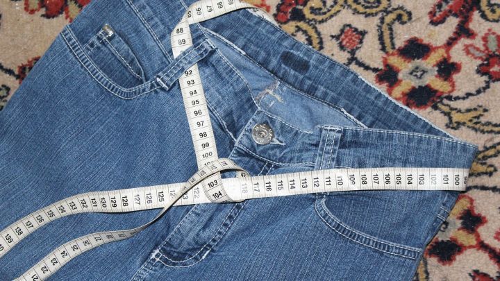 Eine Jeans liegt auf einem dekorativen Teppich, umgeben von einem Ma&szlig;band, das die Ma&szlig;e f&uuml;r das Abnehmen anzeigt.