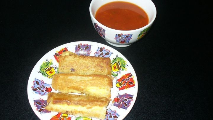 Drei knusprige K&auml;se-Toast-R&ouml;llchen liegen auf einem bunten Teller neben einer Sch&uuml;ssel mit Tomatensuppe.