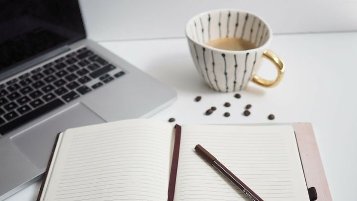 Ein offenes Notizbuch mit einem Stift darauf liegt neben einer Tasse Kaffee und einem Laptop in einem hellen Arbeitsbereich.
