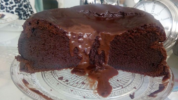 Ein saftiger Schokoladenkuchen steht auf einem eleganten Glasstand, w&auml;hrend ein St&uuml;ck herausgeschnitten ist und Schokolade austritt.