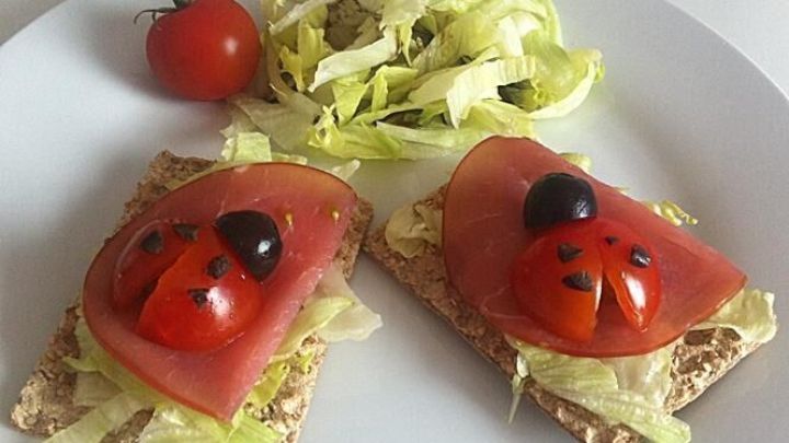 Zwei Brotscheiben sind mit Salat und Tomaten belegt, die wie Marienk&auml;fer aussehen, umgeben von einem kleinen Salat und einer Tomate.