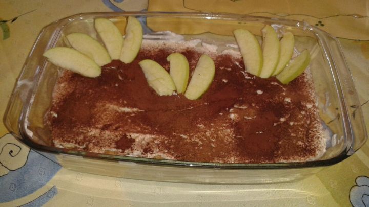 Ein Apfeltiramisu in einer Glasform mit Kakaopulver bestreut, dekoriert mit Apfelscheiben auf einem bunt gemusterten Tischtuch.