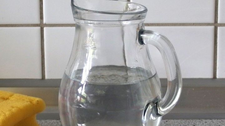 Eine Glas Umsch&uuml;ttkanne f&uuml;llt sich mit Wasser auf einer K&uuml;chentheke, neben einem gelben Schwamm und Fliesen.
