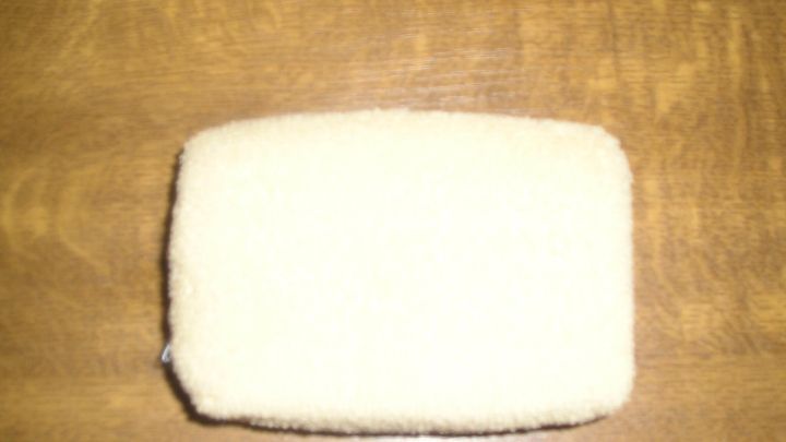 Ein rechteckiger, cremefarbener Schwamm liegt unbewegt auf einer h&ouml;lzernen Tischoberfl&auml;che.