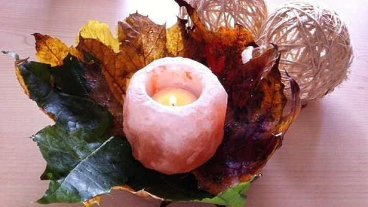 Eine Dekoschale aus buntem Herbstlaub umgibt eine Kerze, die sanft leuchtet, auf einem hellen Tisch.