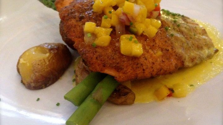 Ein gegrillter Fisch liegt auf Spargel und Kartoffeln, garniert mit einer fruchtigen Salsa, in einem eleganten Restaurant.
