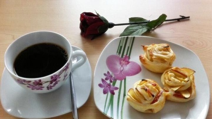 Eine Tasse Kaffee steht neben einem Teller mit Apfel-Bl&auml;tterteig-Rosen, dekoriert mit einer roten Rose auf dem Tisch.