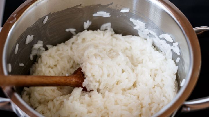 Ein Topf mit gekochtem Reis steht auf dem Herd, w&auml;hrend ein Holzl&ouml;ffel den Reis lockert. Der Kontext ist eine K&uuml;che.