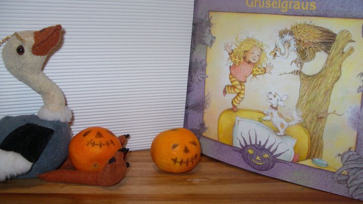 Eine Kuscheltiergans steht neben dem Kinderbuch &bdquo;Fledermaus und Gruselgraus&ldquo; auf einem Holztisch, umgeben von K&uuml;rbisfiguren.