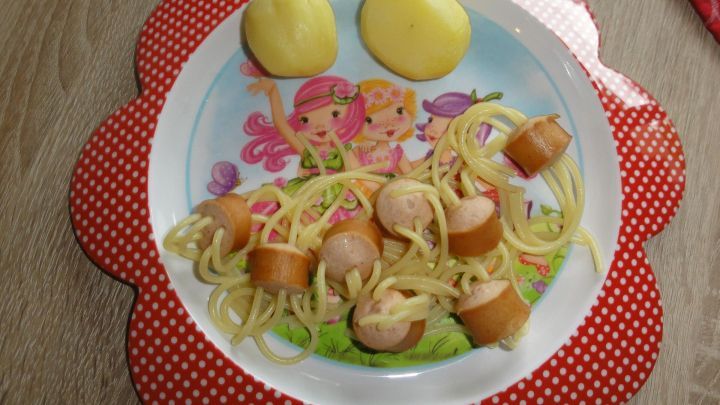 Teller mit "Spinnen"-Gericht: Nudeln durchstechen W&uuml;rstchen, dekoriert mit Kartoffeln. Bunter Hintergrund mit Figuren.