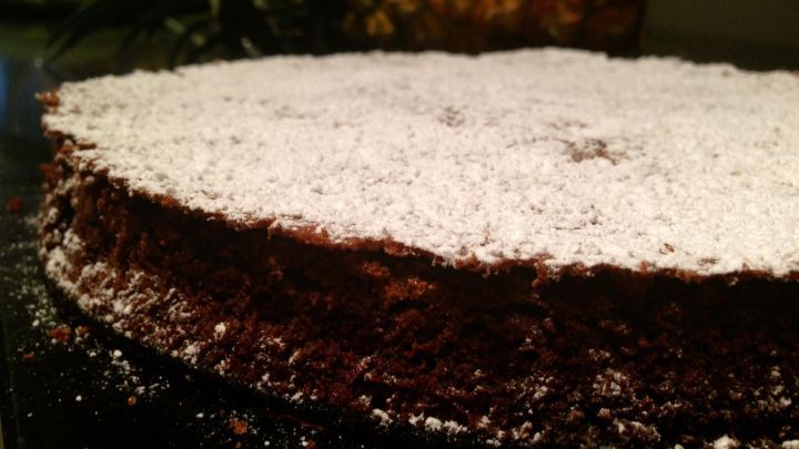 Ein Schokoladenkuchen liegt auf einer dunklen Fl&auml;che, leicht mit Puderzucker best&auml;ubt, vor einem unscharfen Hintergrund.