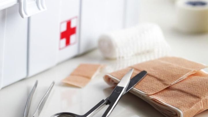 Eine Schere und Pinzette liegen vor Verb&auml;nden und Pflastern. Im Hintergrund ist eine Hausapotheke mit rotem Kreuz zu sehen.