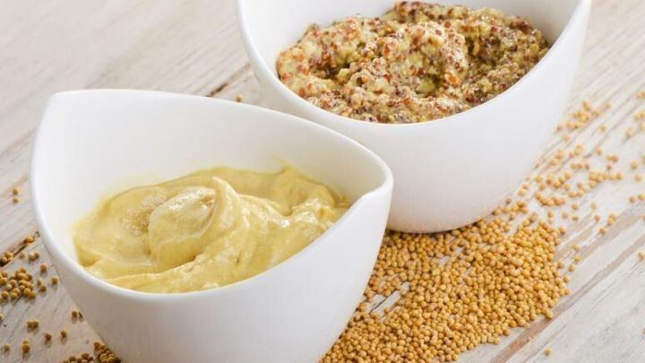 Zwei wei&szlig;e Schalen mit selbstgemachtem Senf, einer glatt und einer grob, stehen auf einem Holztisch umgeben von Senfk&ouml;rnern.