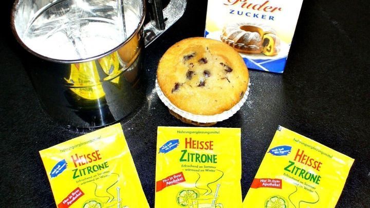 Eine Tasse mit Wasser steht neben drei Packungen "Hei&szlig;e Zitrone" und einem Cookie auf einem dunklen Tisch.